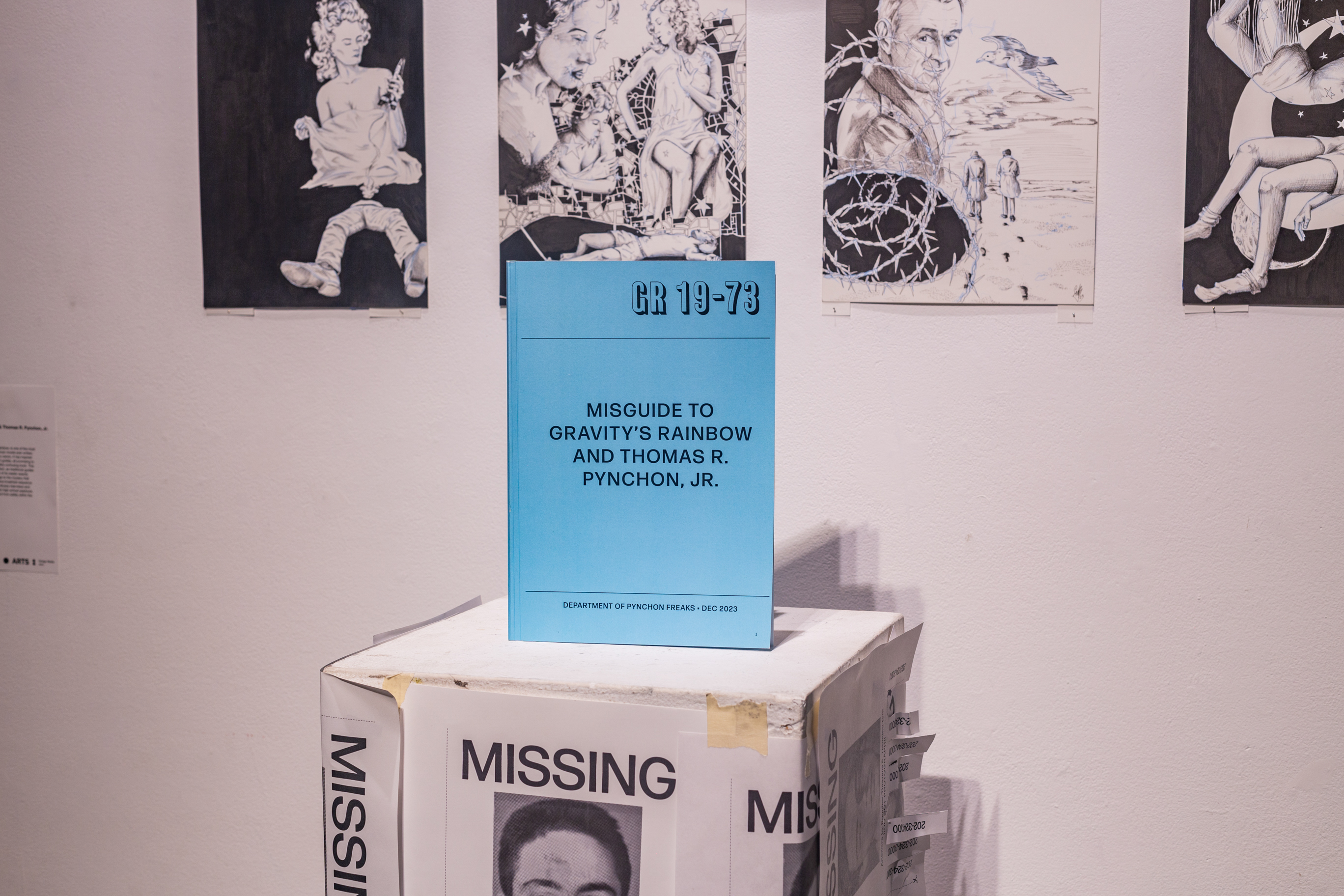 A podium with multiple fabricated missing person posters. On the podium is a book standing upright with the title reading Misguide to Gravity's Rainbow and Thomas R Pynchon Jr.